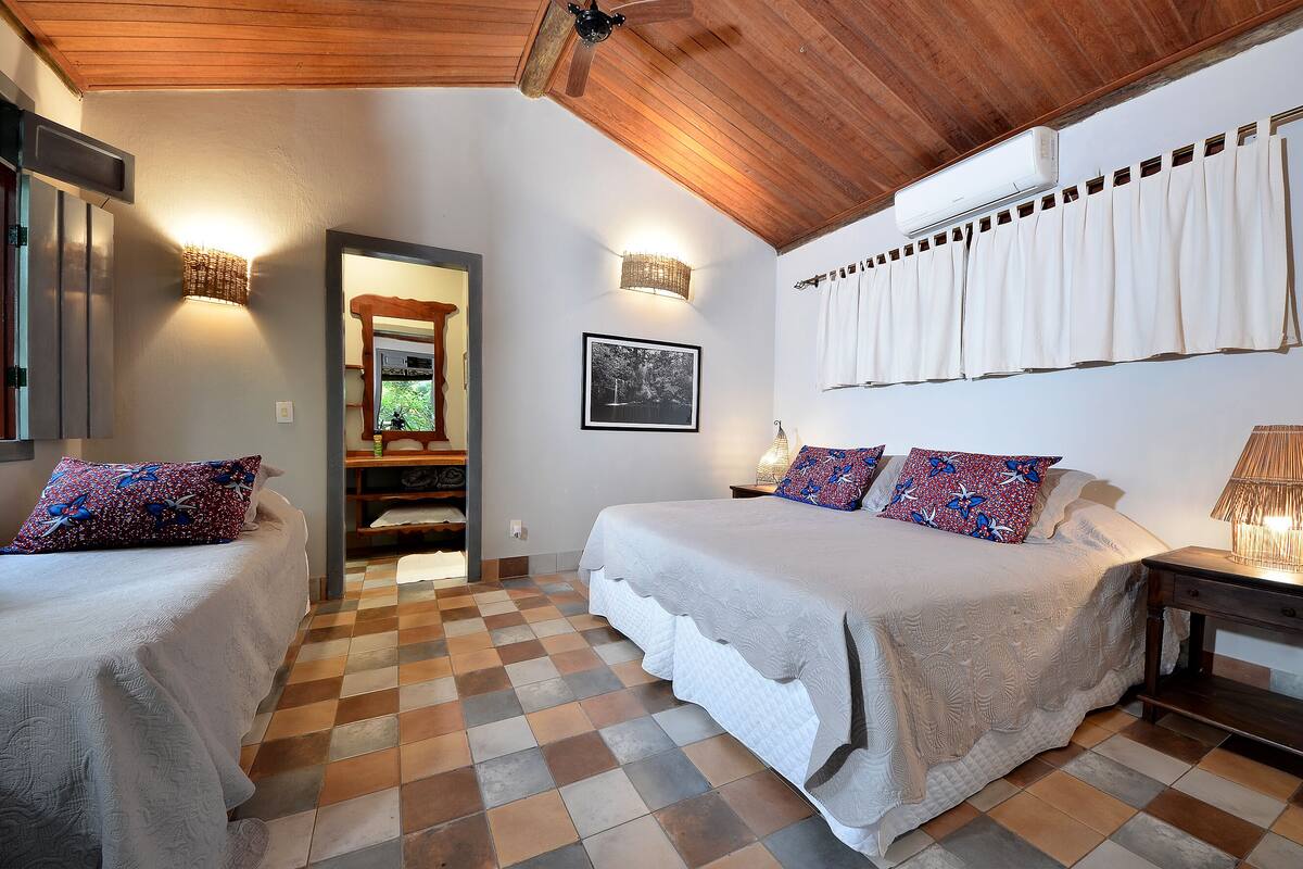 A bedroom with two beds, checkered floor tiles, a wooden ceiling, and a window with white curtains.