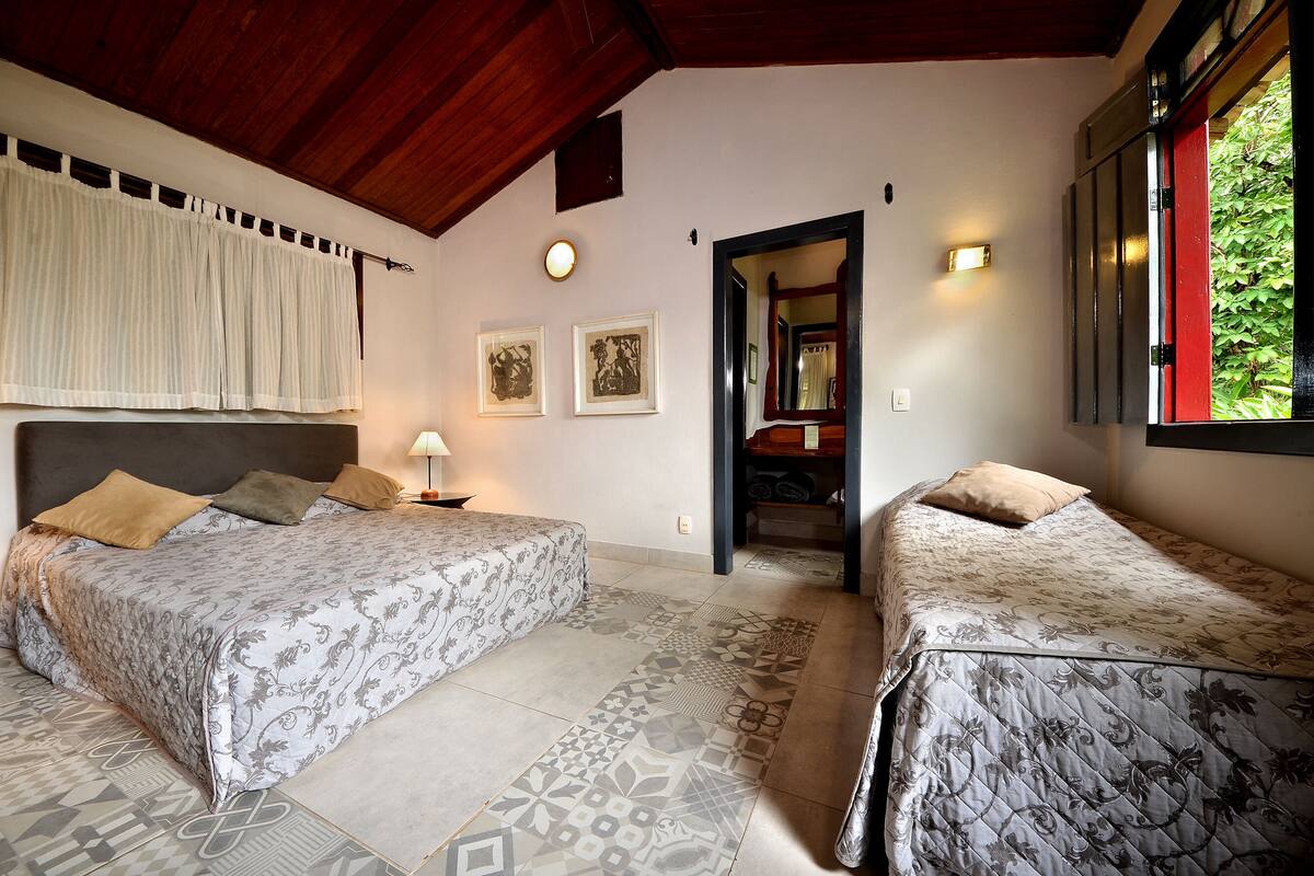 A bedroom with two beds, a wooden ceiling, a window with a red frame, and a door leading to another room.
