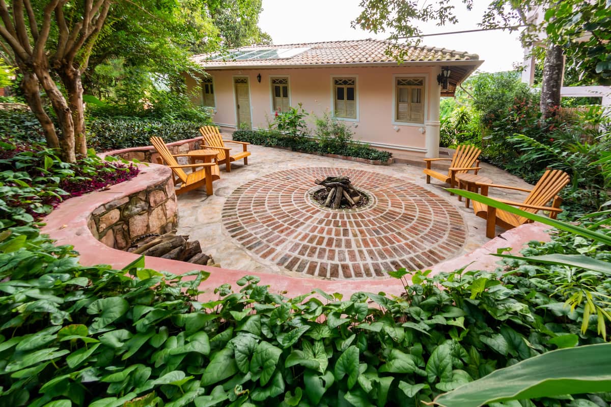 A patio with a circular fire pit surrounded by wooden chairs, a stone wall, and lush greenery.
