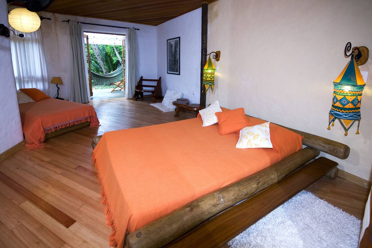 A room with two beds, wooden flooring, a hammock, and a view of greenery outside.