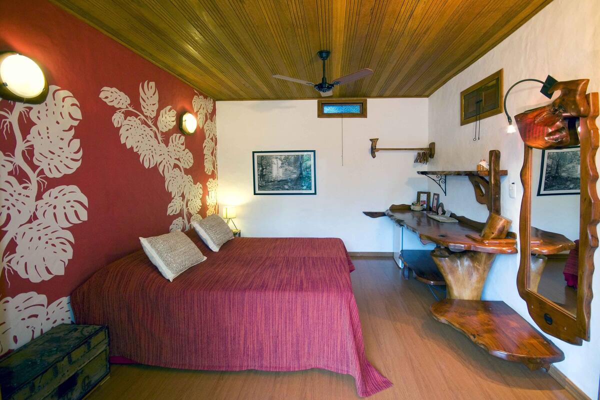A bedroom with a wooden ceiling, a bed with a red bedspread, a wooden nightstand, a wall-mounted mirror, and a framed picture on the wall.
