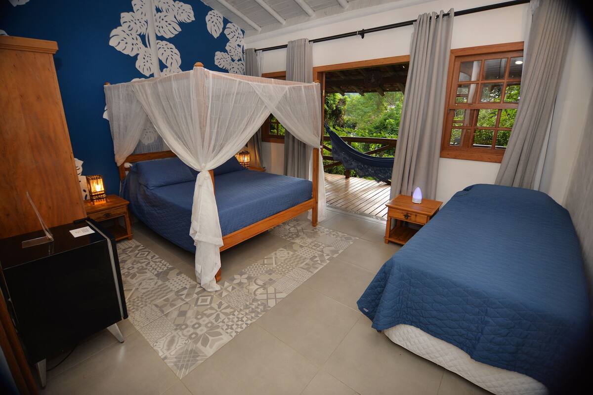A bedroom with a bed under a canopy, a second bed, a nightstand, a wooden wardrobe, and a view of greenery through the window.