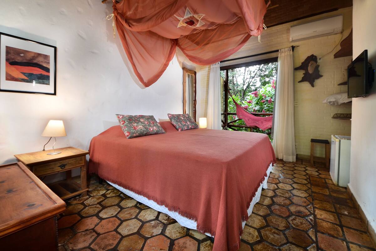 A bedroom with a bed, a nightstand, a lamp, a TV, and a view of greenery outside.