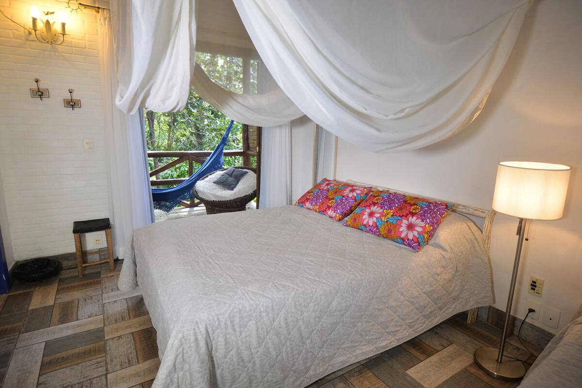 A bedroom with a bed, a nightstand, a lamp, a window with curtains, and a view of greenery outside.