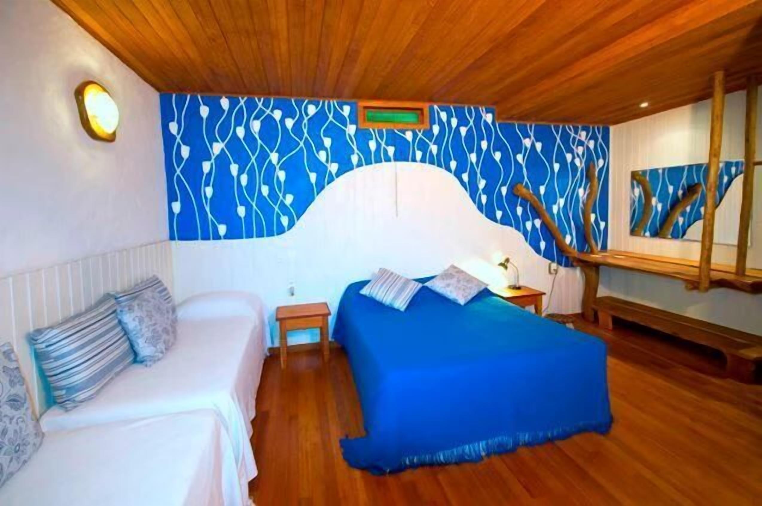 A bedroom with a wooden ceiling, a blue and white patterned wall, a bed with a blue bedspread, a bedside table, and a nightstand.