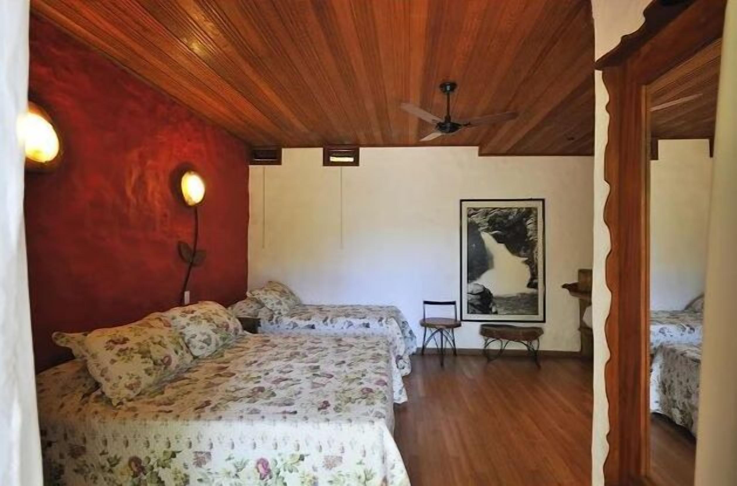 A room with two beds, wooden ceiling, a ceiling fan, a framed picture on the wall, and a small table with a chair.