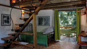 Une pièce rustique avec un escalier en bois, un hamac, un lit recouvert d’une couverture verte et une table en bois.