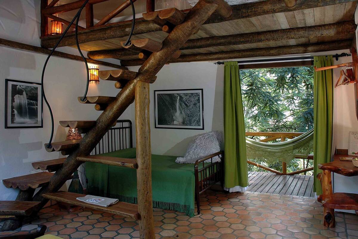 A rustic room with a wooden staircase, a hammock, a bed with a green blanket, and a wooden table.