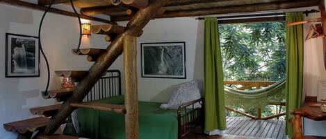 A rustic room with a wooden staircase, a hammock, a bed with a green blanket, and a wooden table.