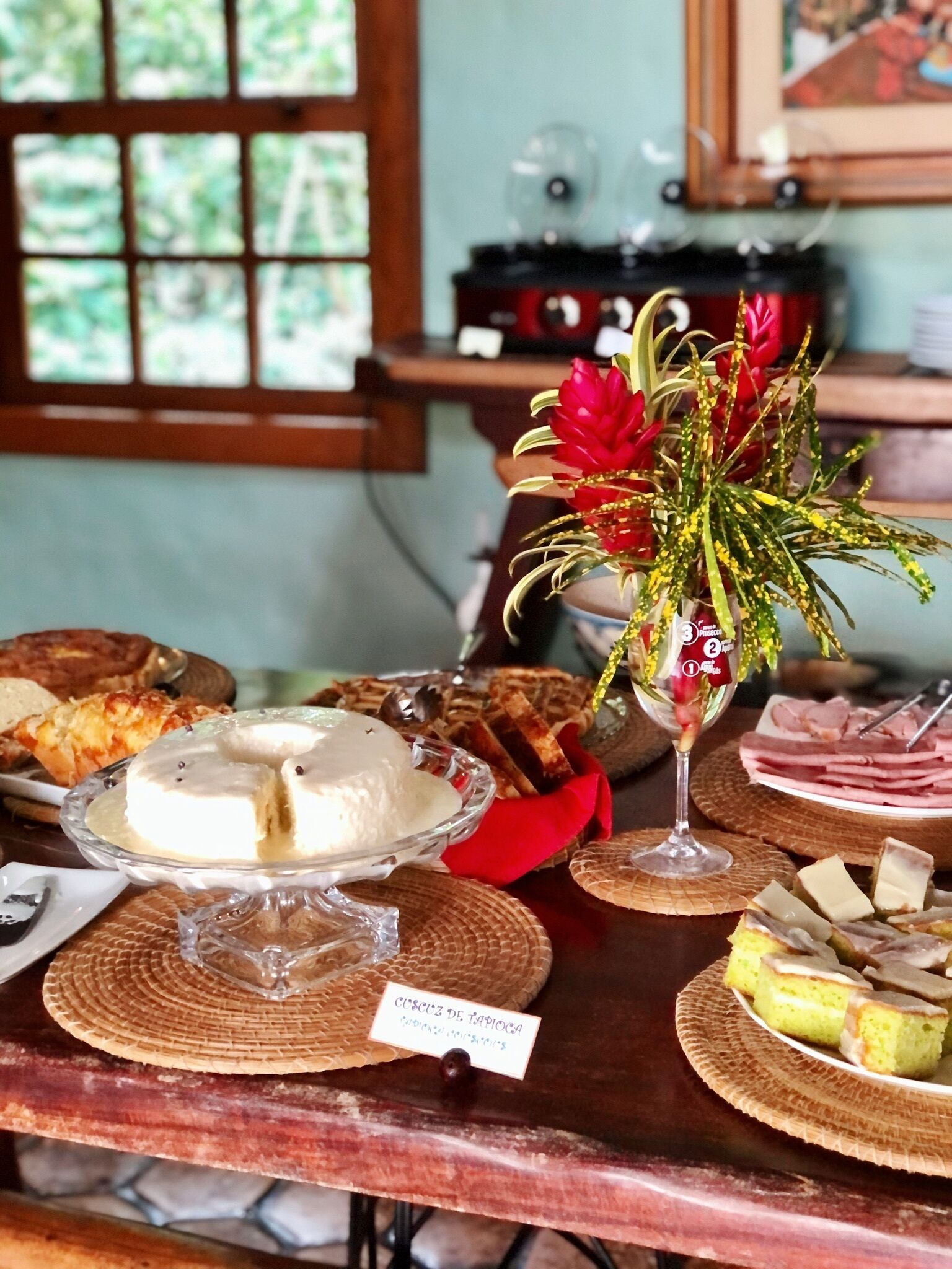 A buffet table with various food items, including a cake labeled 'Cuscillo de Tapioca', a glass bowl of white food, and a red flower arrangement.