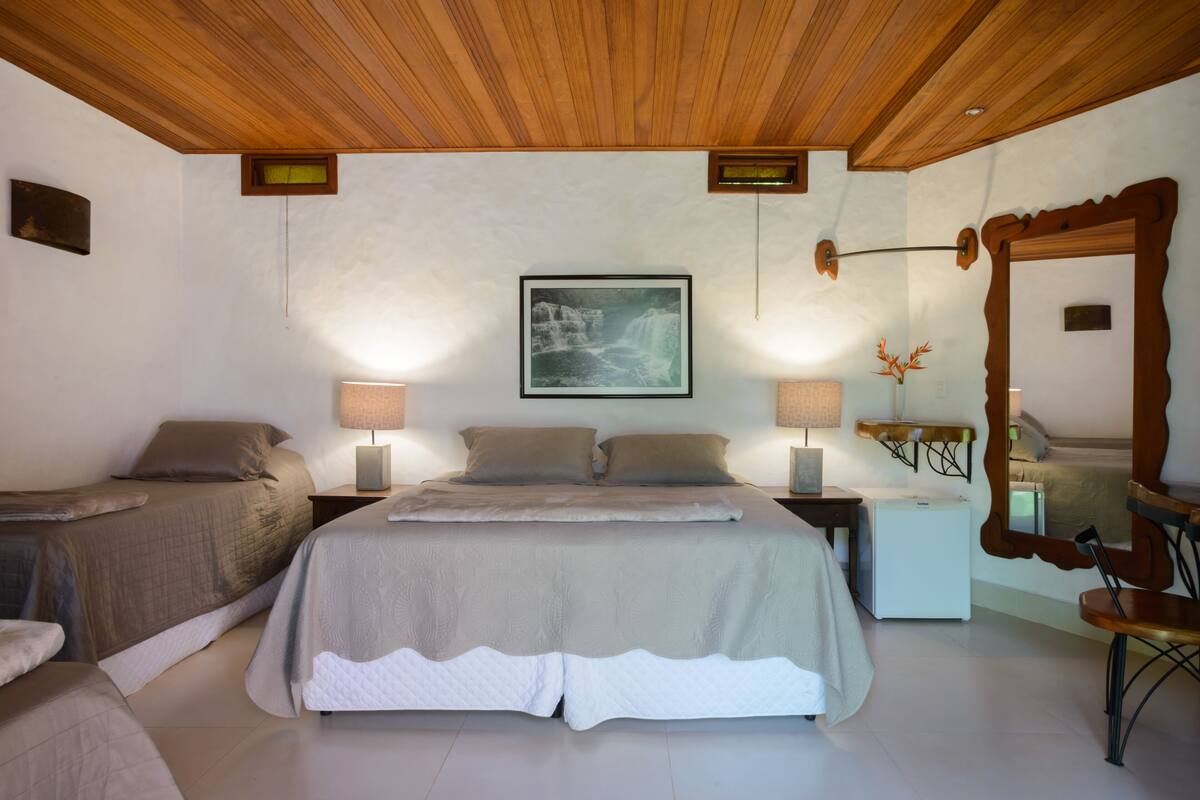 A bedroom with a wooden ceiling, a bed with a white and beige bedspread, two bedside tables with lamps, a framed picture on the wall, and a mirror with an ornate frame.