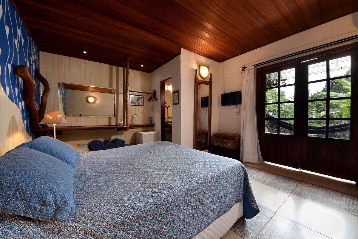 A bedroom with a large bed, wooden ceiling, a large window with a view, a mirror, and a TV.