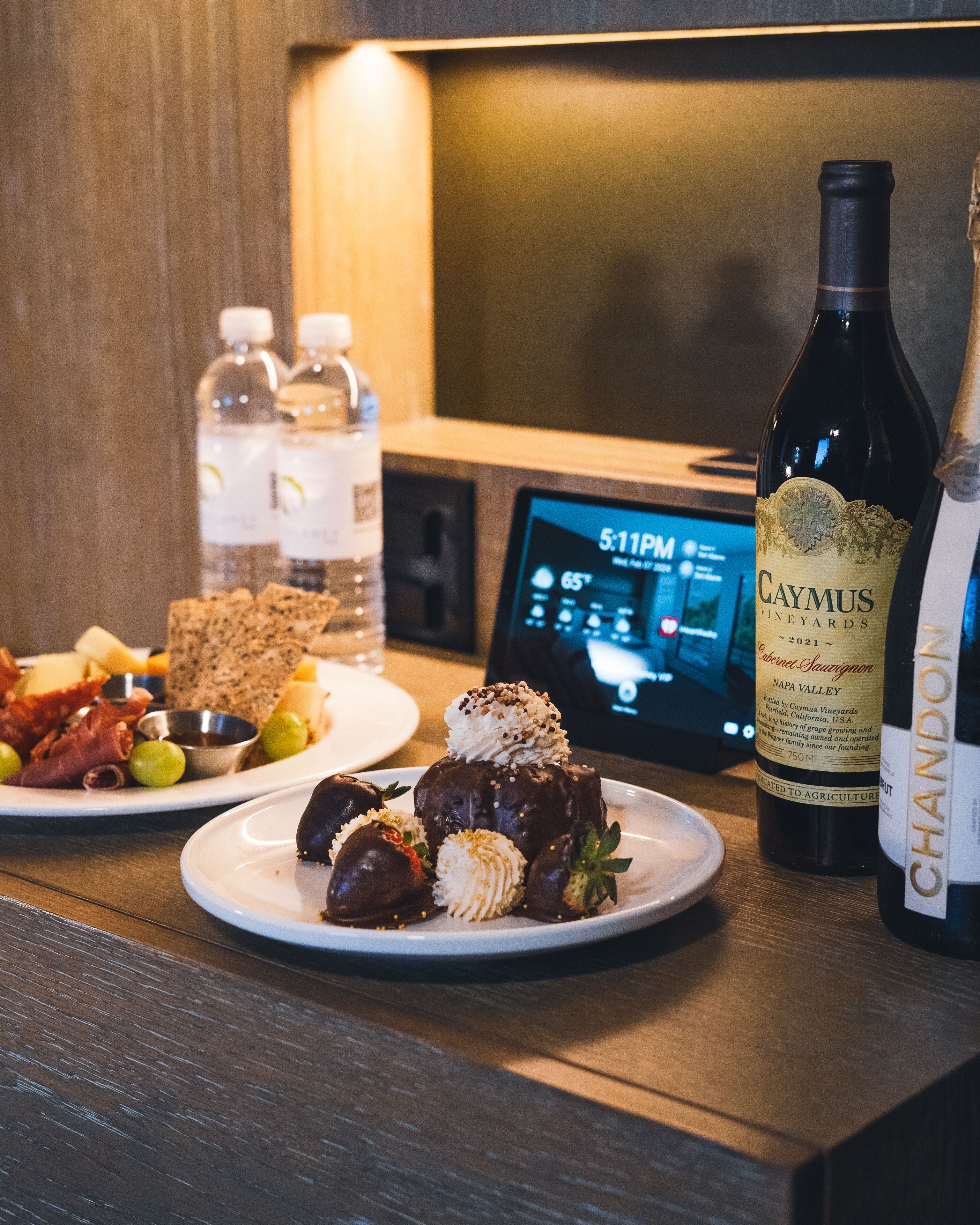 Una mesa con una botella de Caymus Napa Valley Cabernet Sauvignon, un plato de fresas cubiertas de chocolate, y una selección de quesos y embutidos.