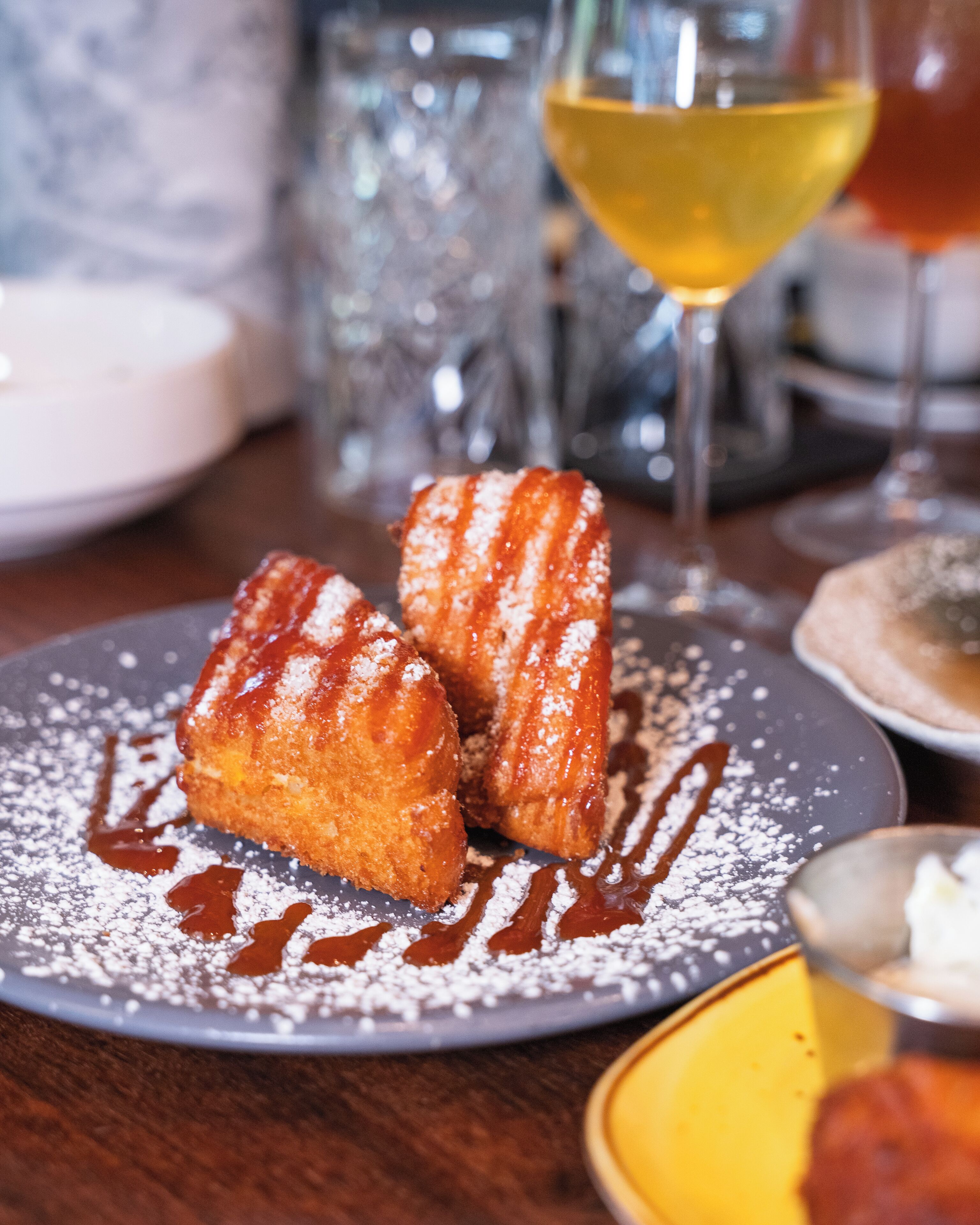 A plate with two fritters, a glass of yellow drink, and a dessert on a table.