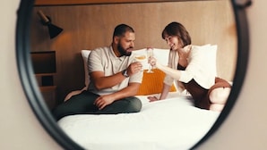 A round mirror reflecting a couple on a bed with a wooden headboard, a lamp, and a small shelf. - The Delaney Hotel (Orlando)