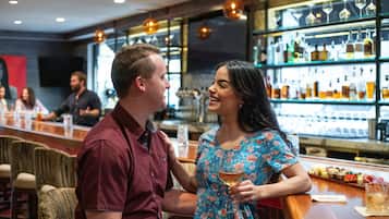 A couple at a bar with a wine glass, seated at a wooden bar counter with a variety of bottles and glasses in the background.