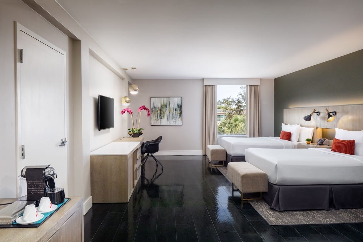 A modern hotel room with a flat-screen TV, a desk with a coffee maker, two beds, a small seating area, and a large window with a view of greenery.