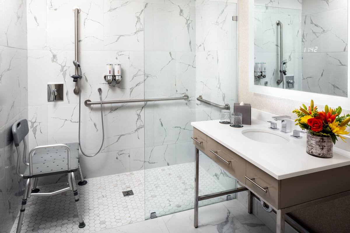 A modern bathroom with a white marble wall, a glass shower enclosure, a white sink with a vase of flowers, and a wheelchair-accessible shower chair.