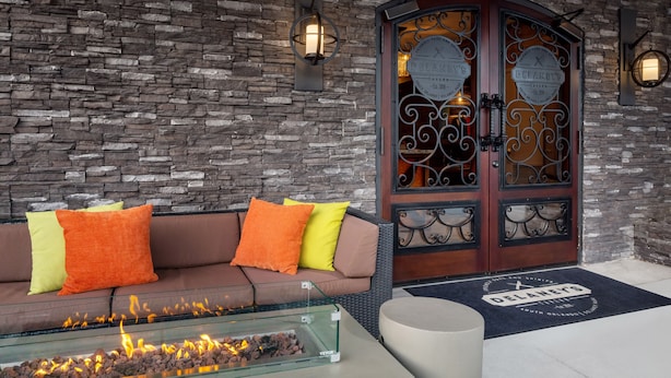 A stone wall, a brown sofa with orange and yellow cushions, a glass table with a fire feature, and a wooden door with decorative glass panels. - The Delaney Hotel (Orlando)