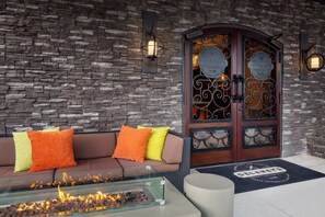 Una pared de piedra, un sofá marrón con cojines naranja y amarillos, una mesa de vidrio con chimenea integrada, y una puerta de madera con paneles de vidrio decorativos.