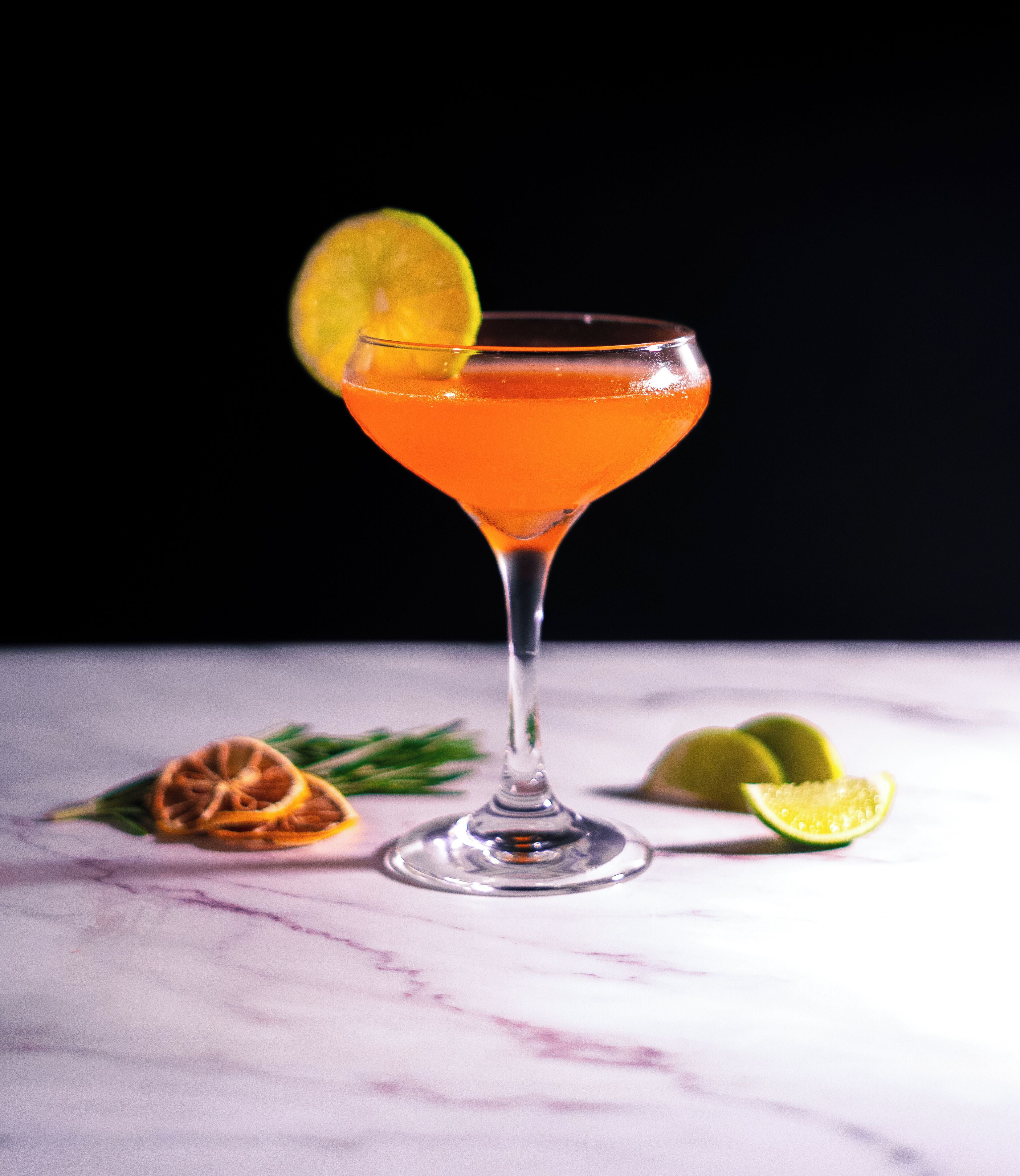 A cocktail in a martini glass with a lime garnish, placed on a marble surface with a black background.