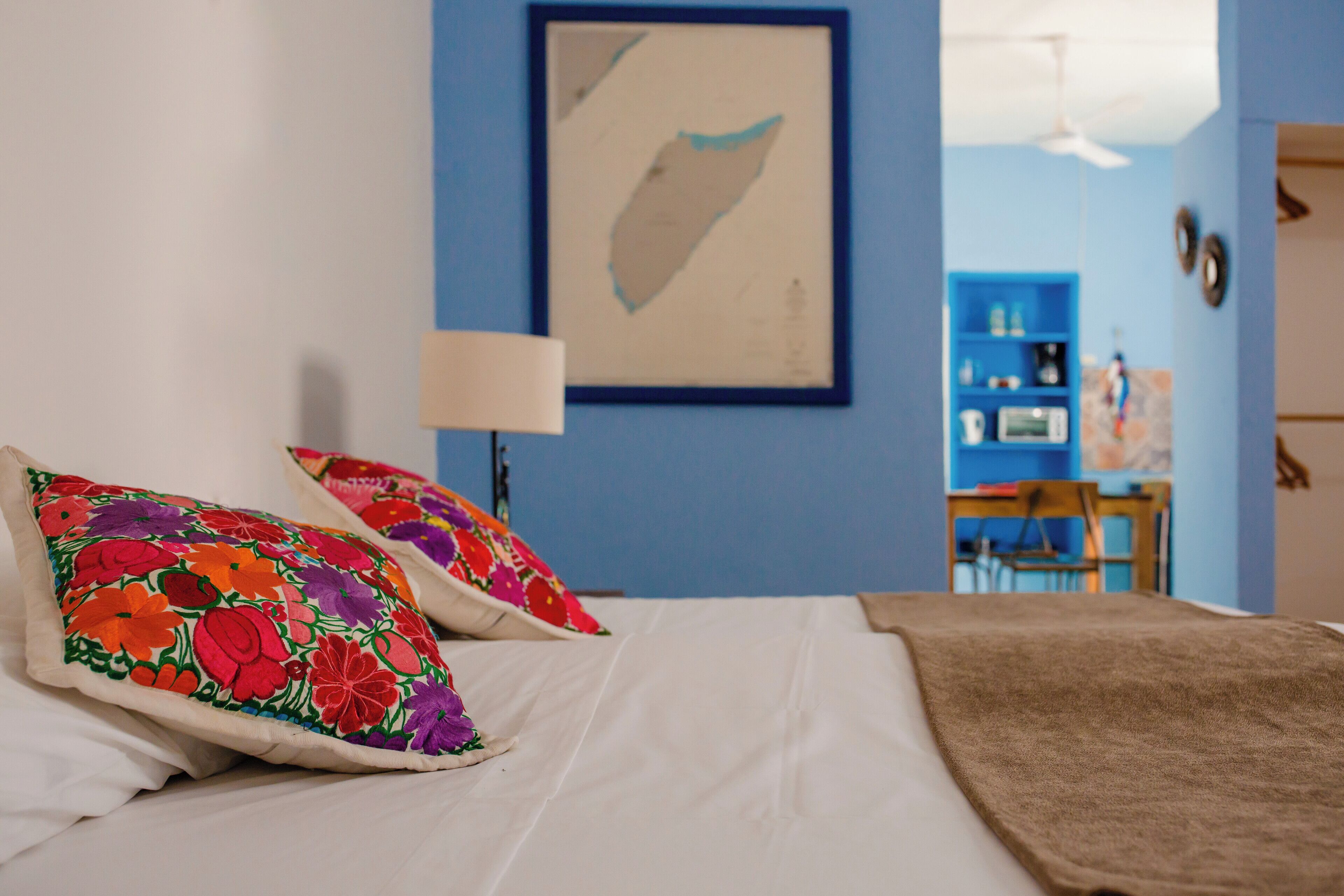 Una cama con almohadones coloridos, una colcha blanca y una pared azul con un cuadro enmarcado.