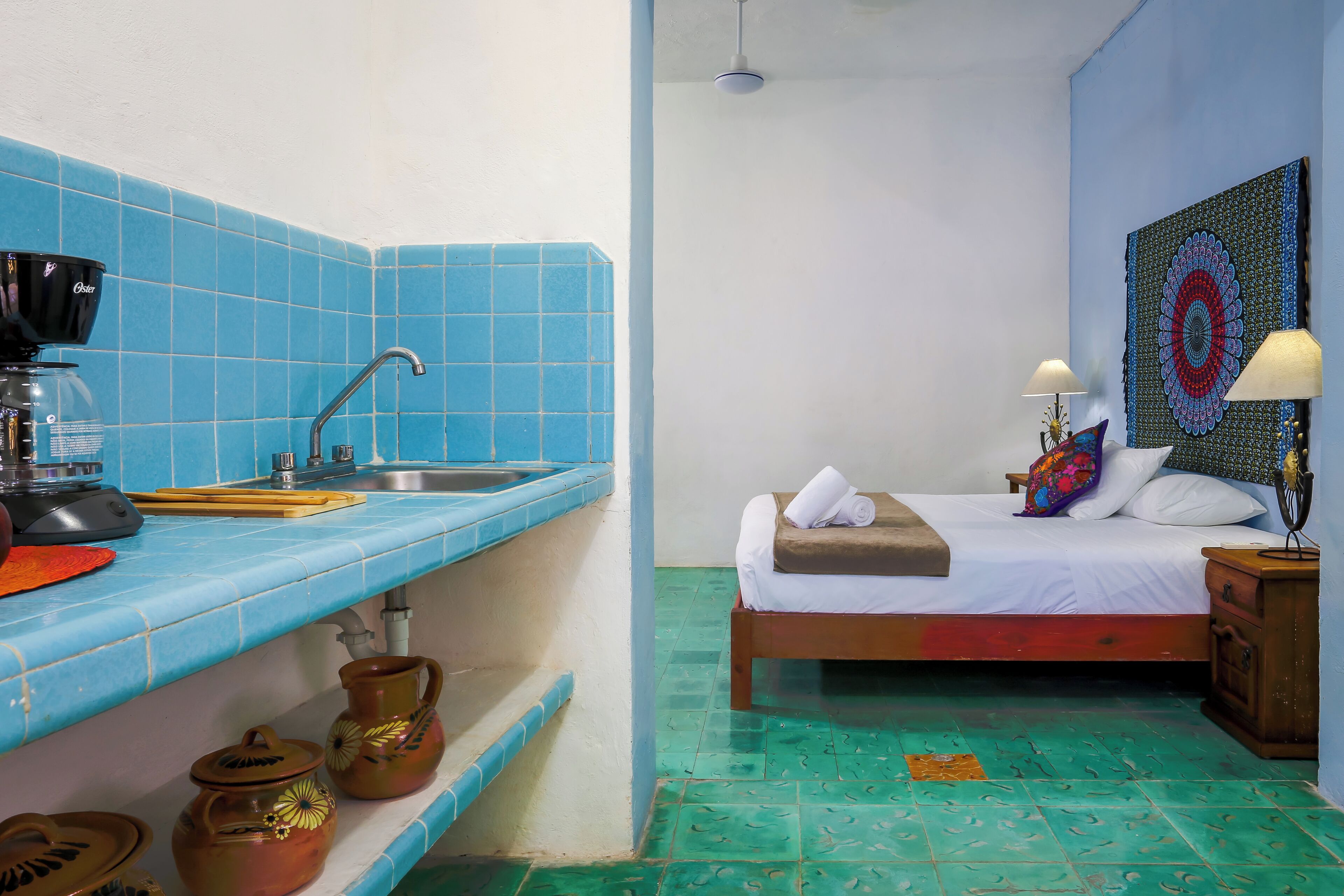A room with a kitchen area featuring a coffee maker, a sink, and a shelf with pottery. There is a bed with a colorful pillow and a nightstand with a lamp.