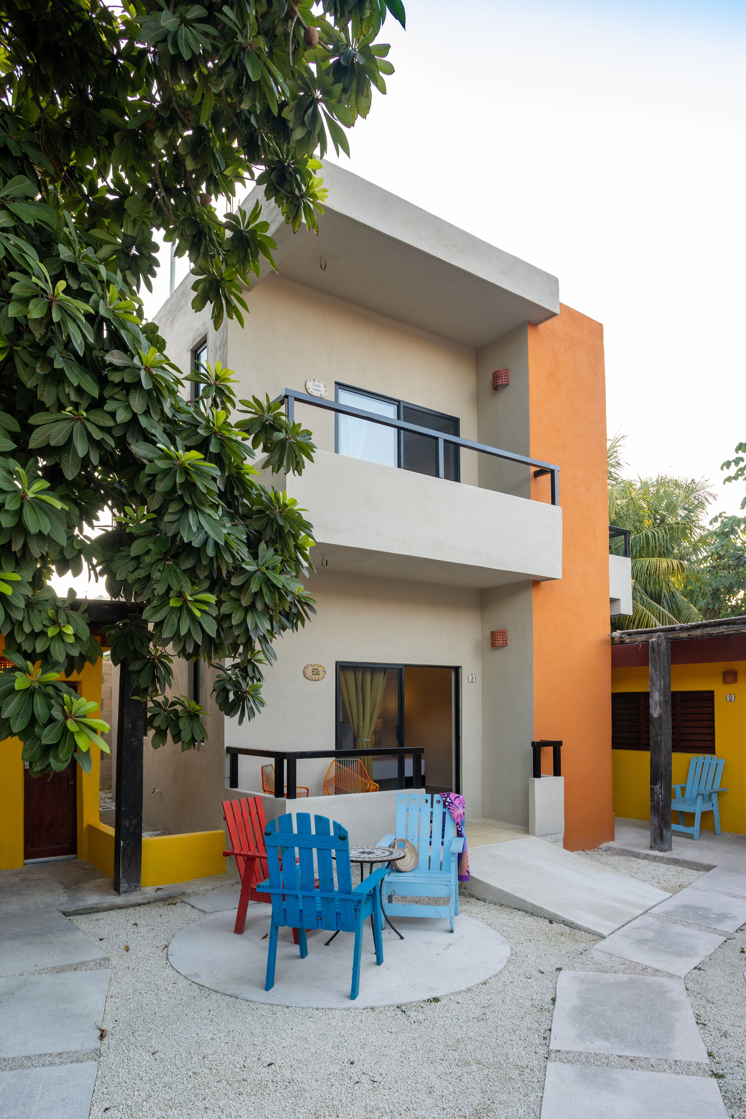 A modern two-story building with a balcony, outdoor seating area with colorful chairs, and a tree in the foreground.