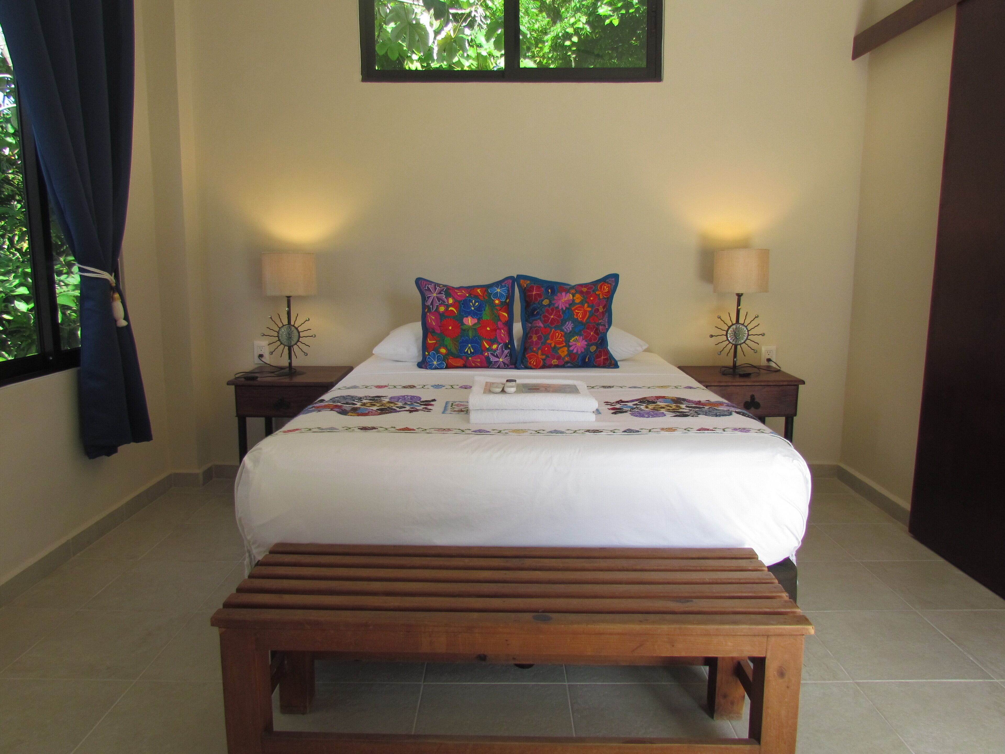 A neatly made bed with a floral quilt, two bedside lamps, and a wooden bench in front.