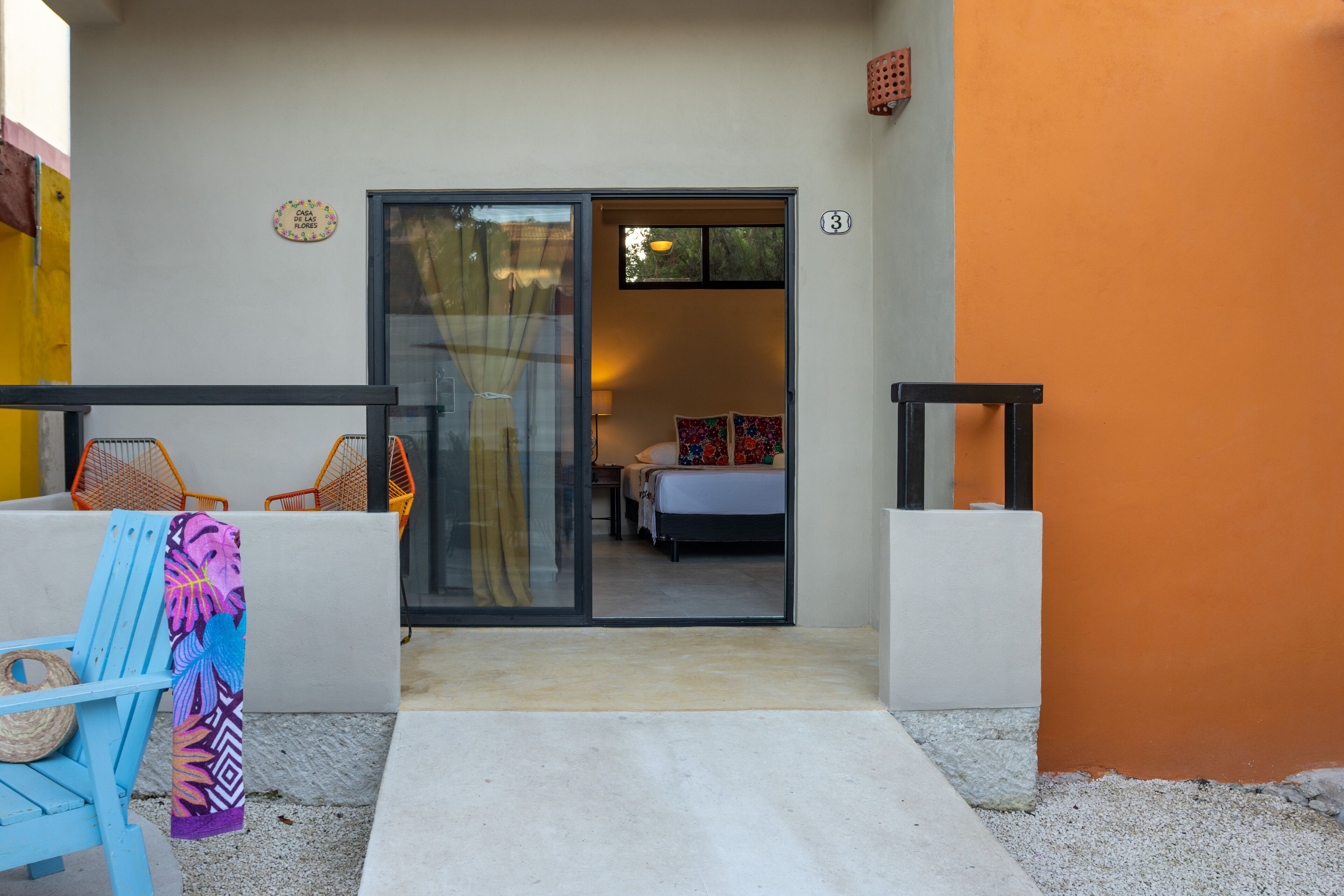 L’entrée d’un hôtel, avec une porte en verre donnant sur une chambre à coucher équipée d’un lit et d’une table de chevet. À l’extérieur, on aperçoit des chaises orange et une serviette à motifs tropicaux est suspendue à l’une d’elles.