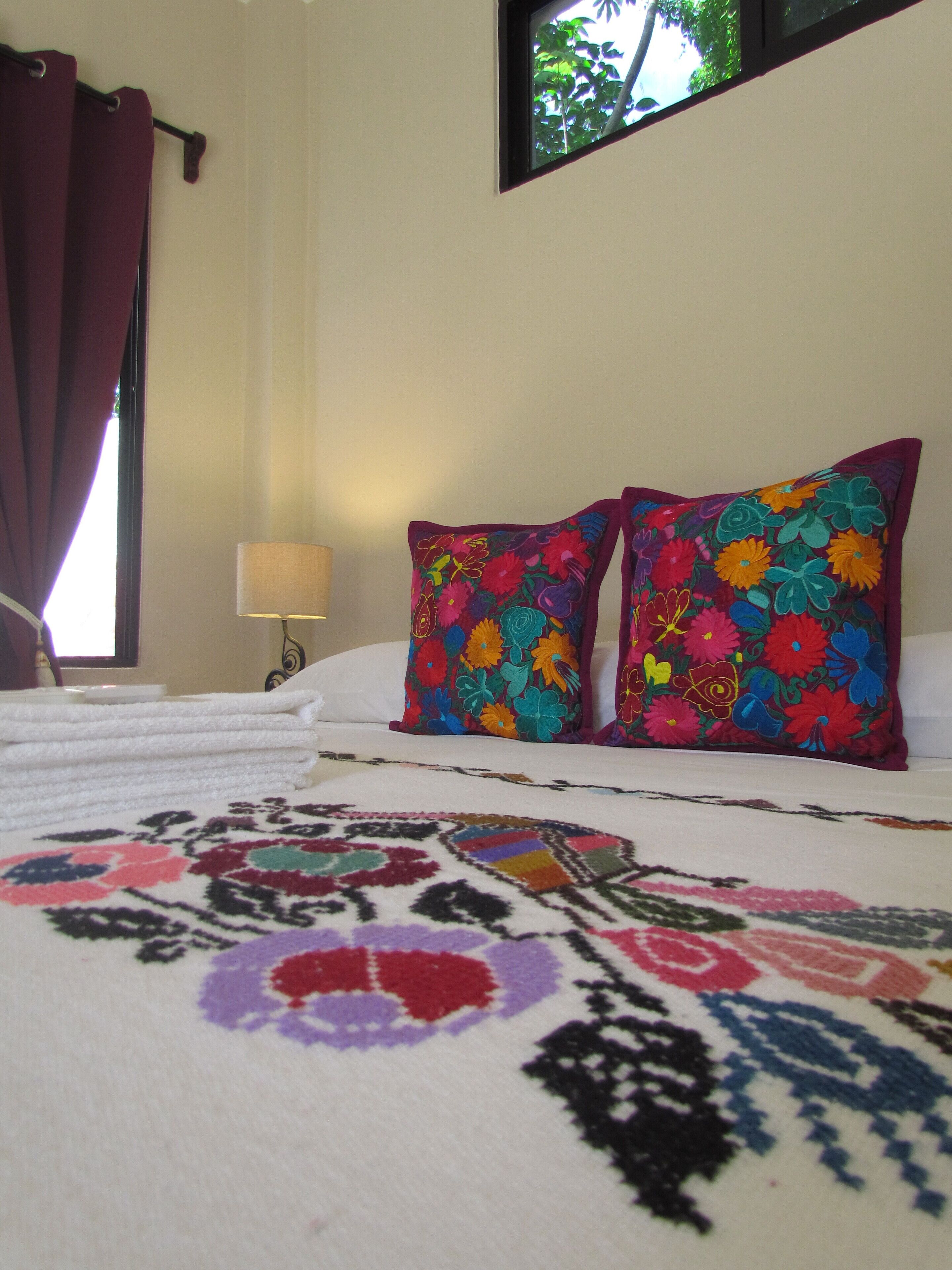 Une chambre d’hôtel avec un lit, deux oreillers colorés, une serviette blanche soigneusement pliée, une lampe et une fenêtre donnant sur des arbres.