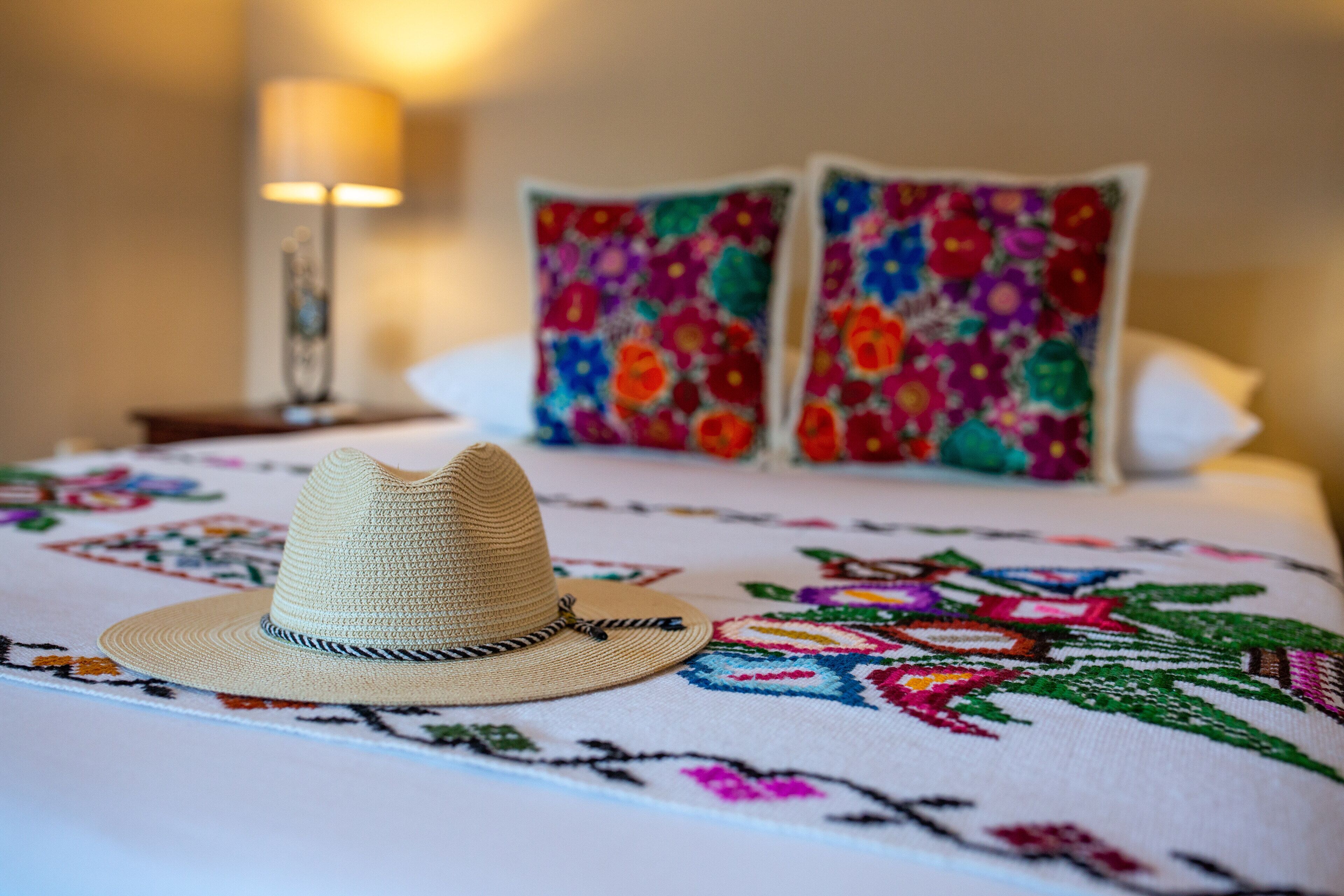 A bed with a colorful embroidered blanket, a straw hat, and two floral pillows.