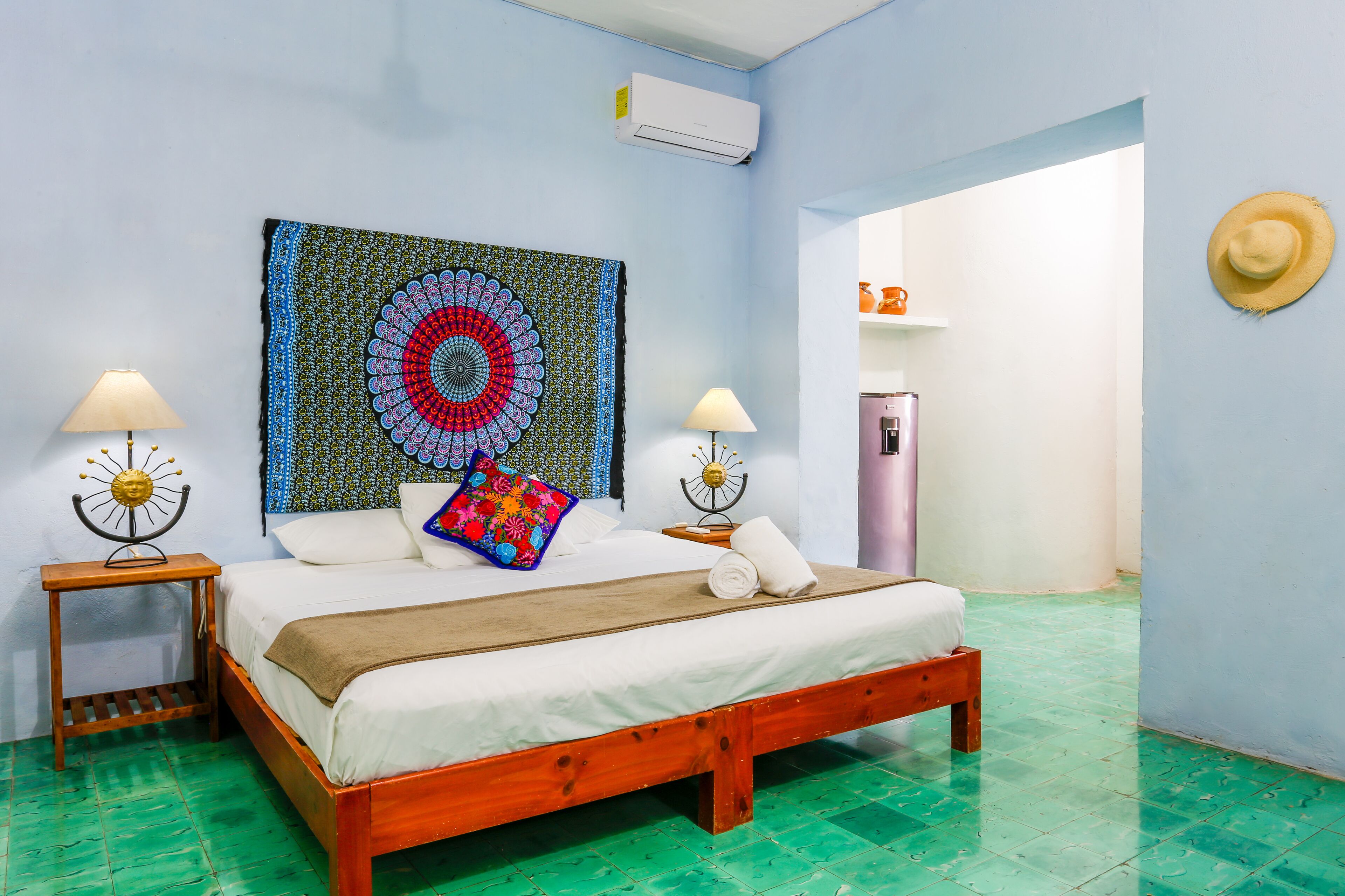 A room with a bed, two nightstands, a wall tapestry, a hat, and a refrigerator.