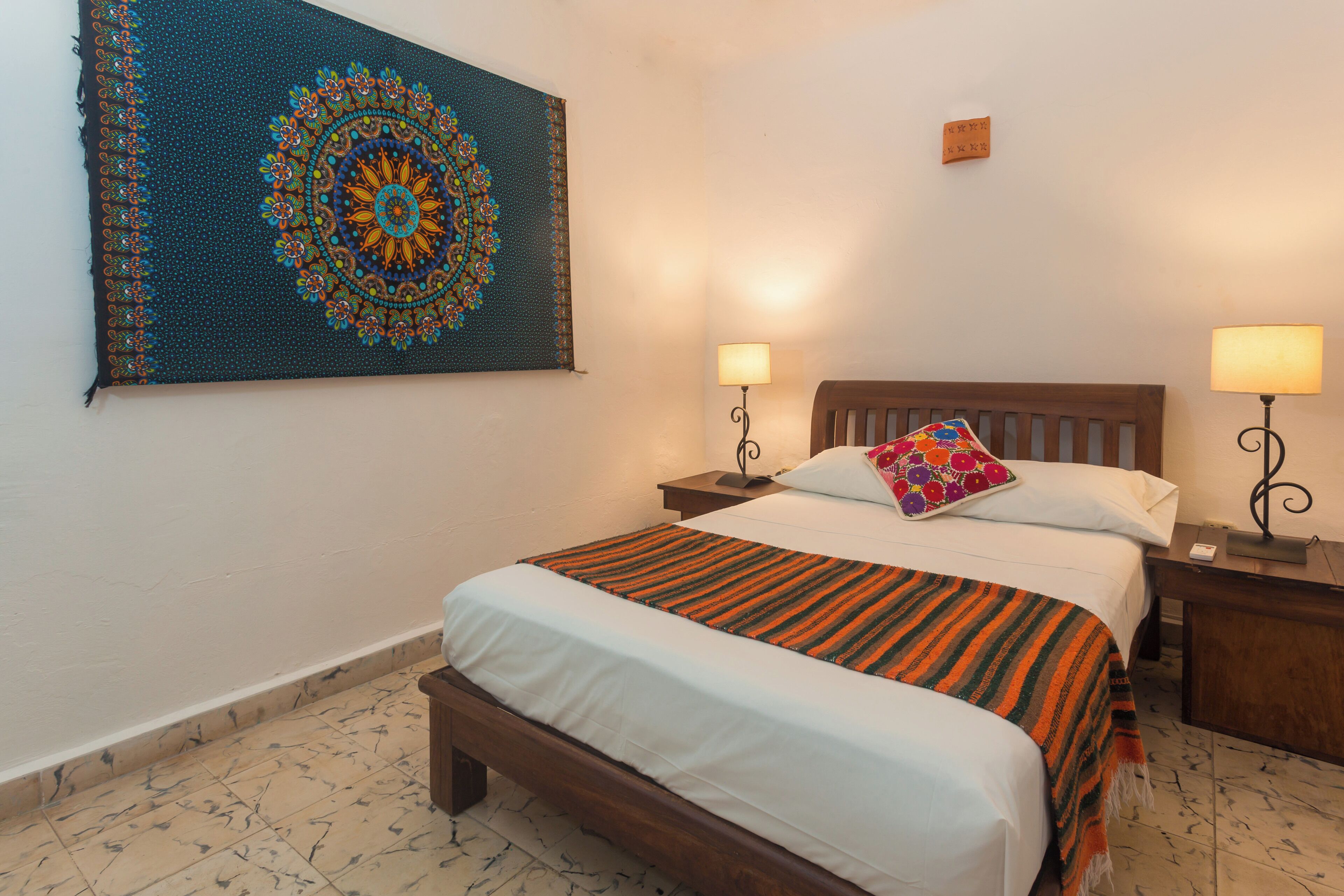 Un lit bien fait, avec une couverture à rayures, deux lampes de chevet, un coussin coloré et une grande tapisserie accrochée au mur.