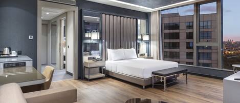 A modern hotel room with a large bed, a desk with a chair, a small table, and a view of the cityscape.