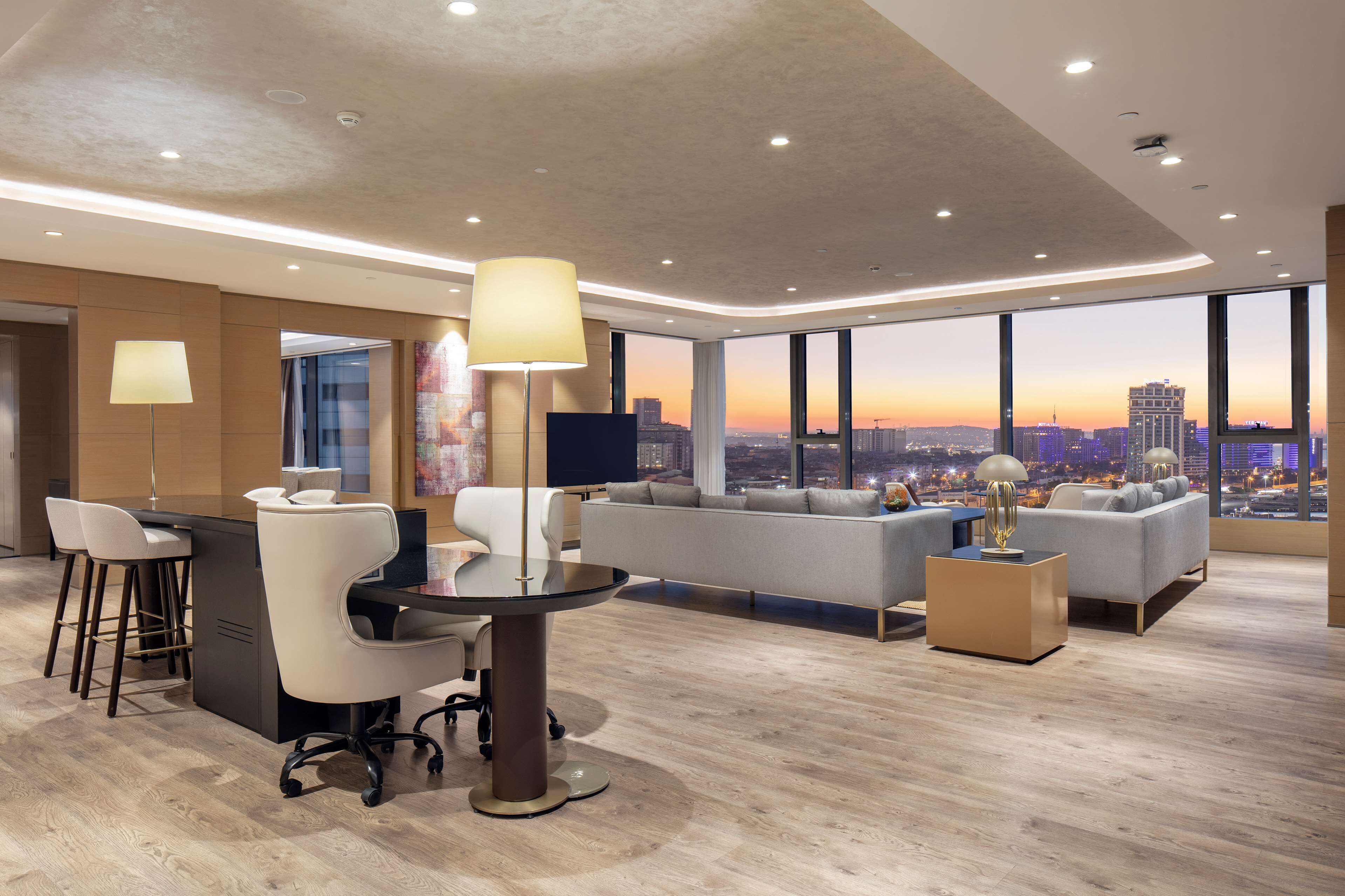 A modern hotel room with a large window offering a city view, a seating area with a sofa and chairs, a dining table with chairs, and a flat-screen TV.