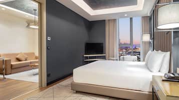 A modern hotel room with a large bed, a TV, a desk with a phone, and a view of the cityscape through large windows.