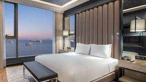 A modern hotel room with a large bed, a bedside table with a lamp, a bench, and a view of the sea with boats. - Hilton Istanbul Bakirkoy (Istanbul)