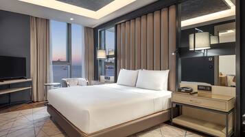 A modern hotel room with a large bed, a TV, a bedside table, a mirror, and a view of the cityscape.