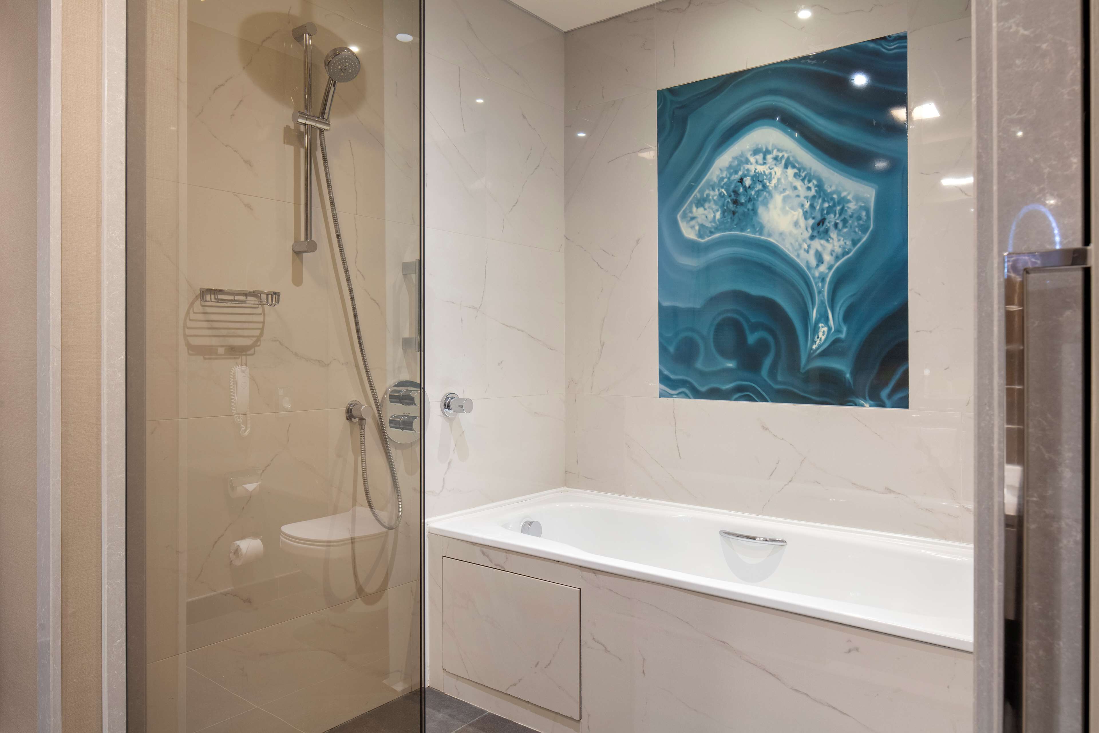 Een moderne badkamer met een douchecabine van glas, een wit bad en een marmeren muur met een blauw abstract schilderij.