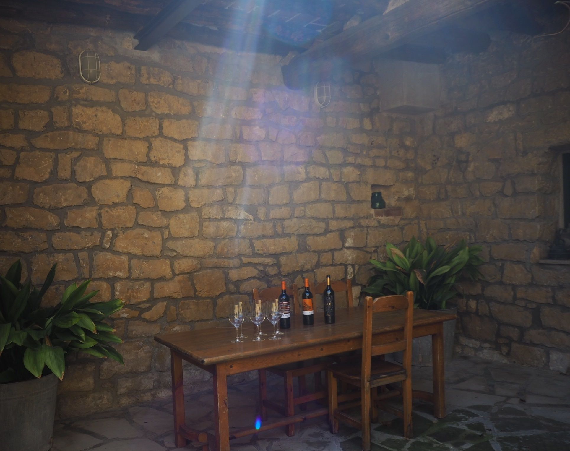 Habitación con paredes de piedra y una mesa de madera dispuesta para comer, con botellas de vino y copas.