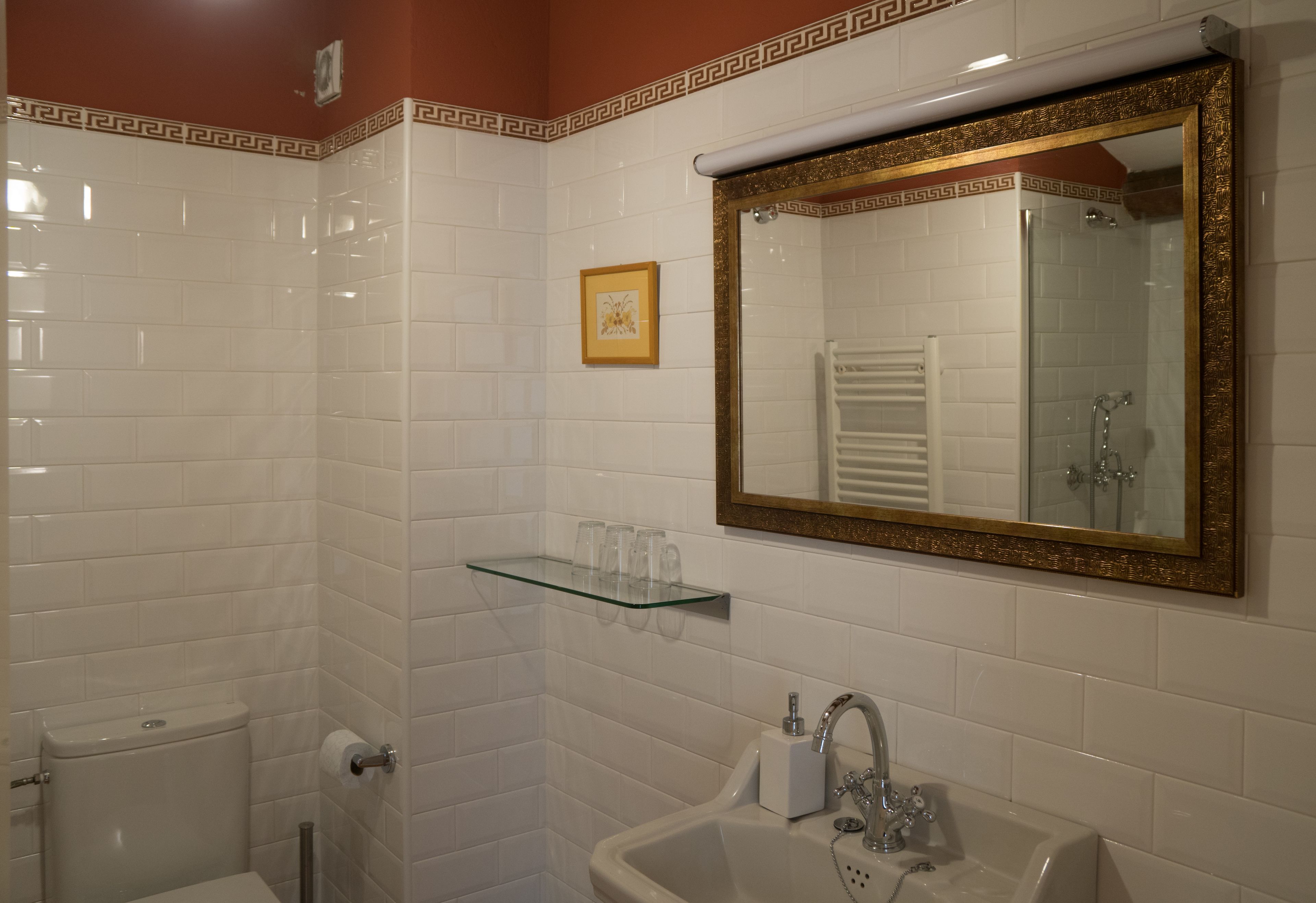Un baño con paredes de azulejos blancos, un espejo con marco dorado, un estante de vidrio con artículos de aseo y un lavamanos blanco.