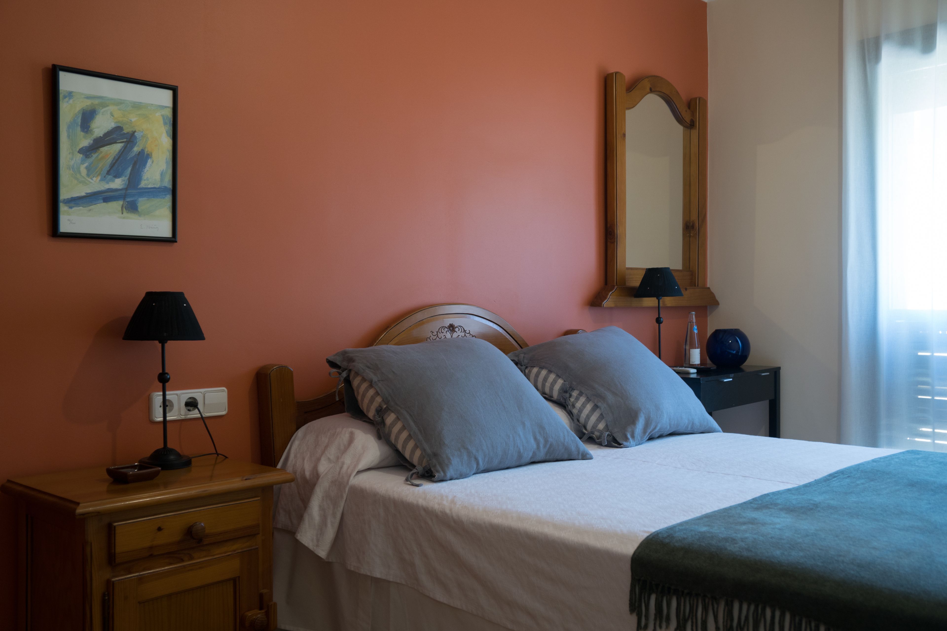 Een slaapkamer met een bed, nachtkastje, lamp, spiegel en een schilderij aan de muur.