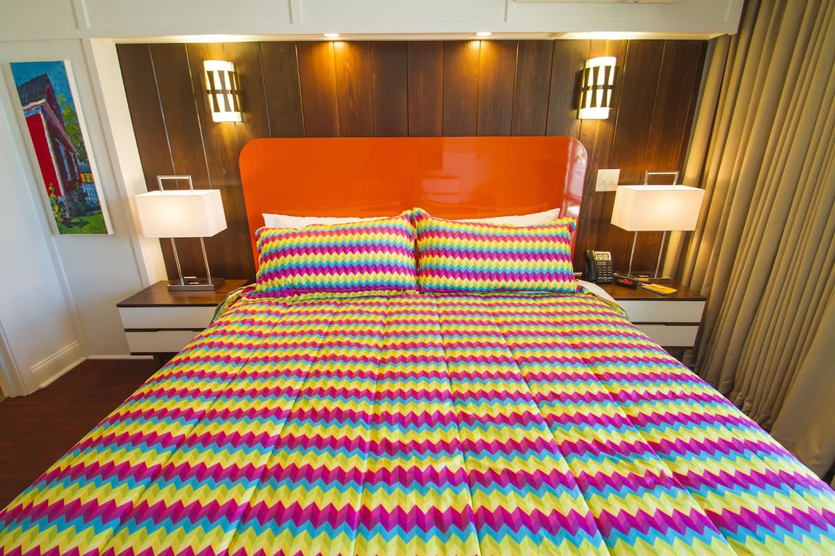 A hotel room with a vibrant zigzag patterned bedspread, wooden headboard, two bedside lamps, and a painting on the wall.