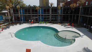 A swimming pool with multiple lanes, surrounded by lounge chairs and a building with large windows in the background. - Mother Earth Motor Lodge (Kinston)