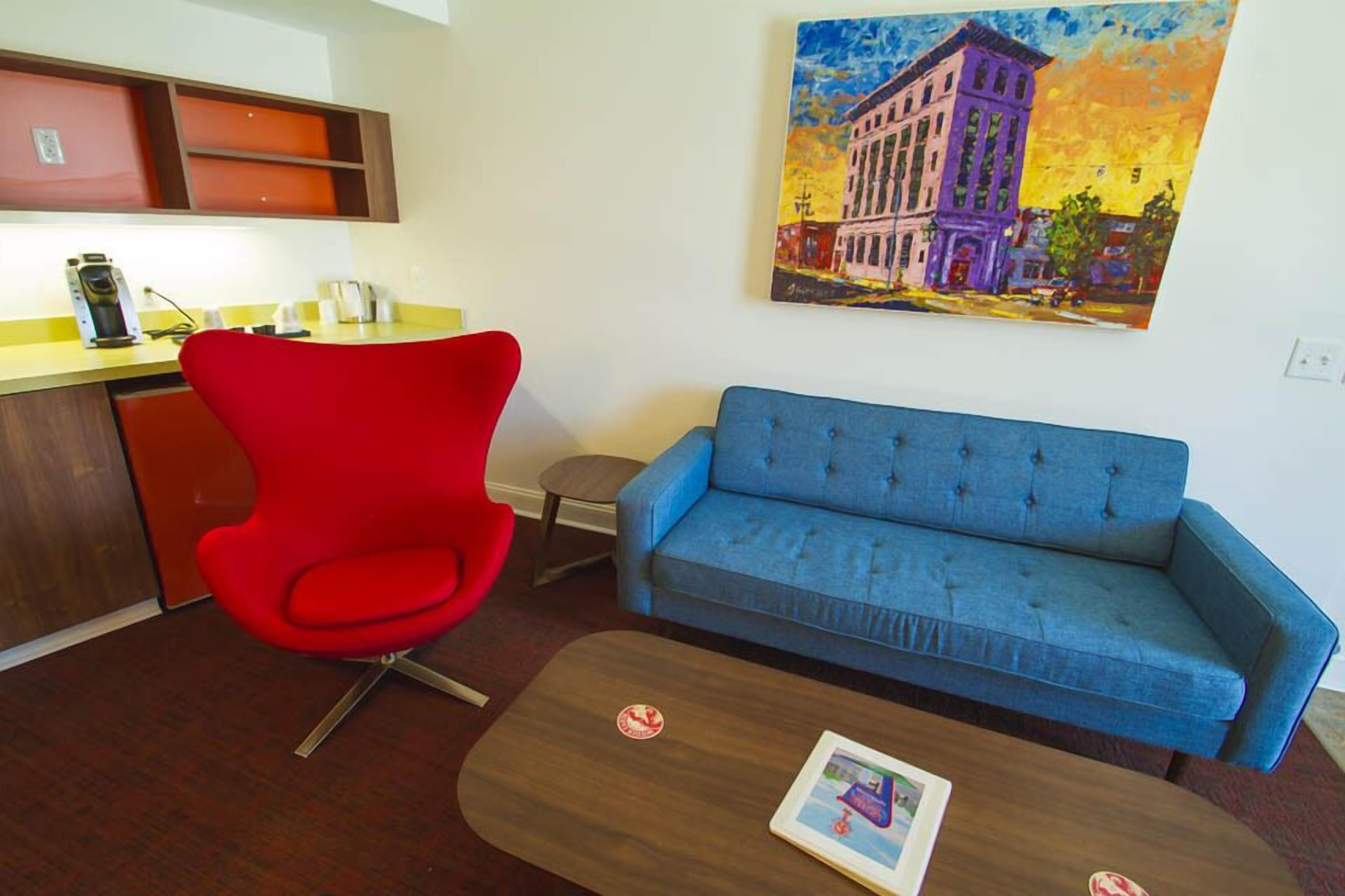 A living room with a red chair, blue sofa, and a coffee table with a book. A painting of a building is on the wall.