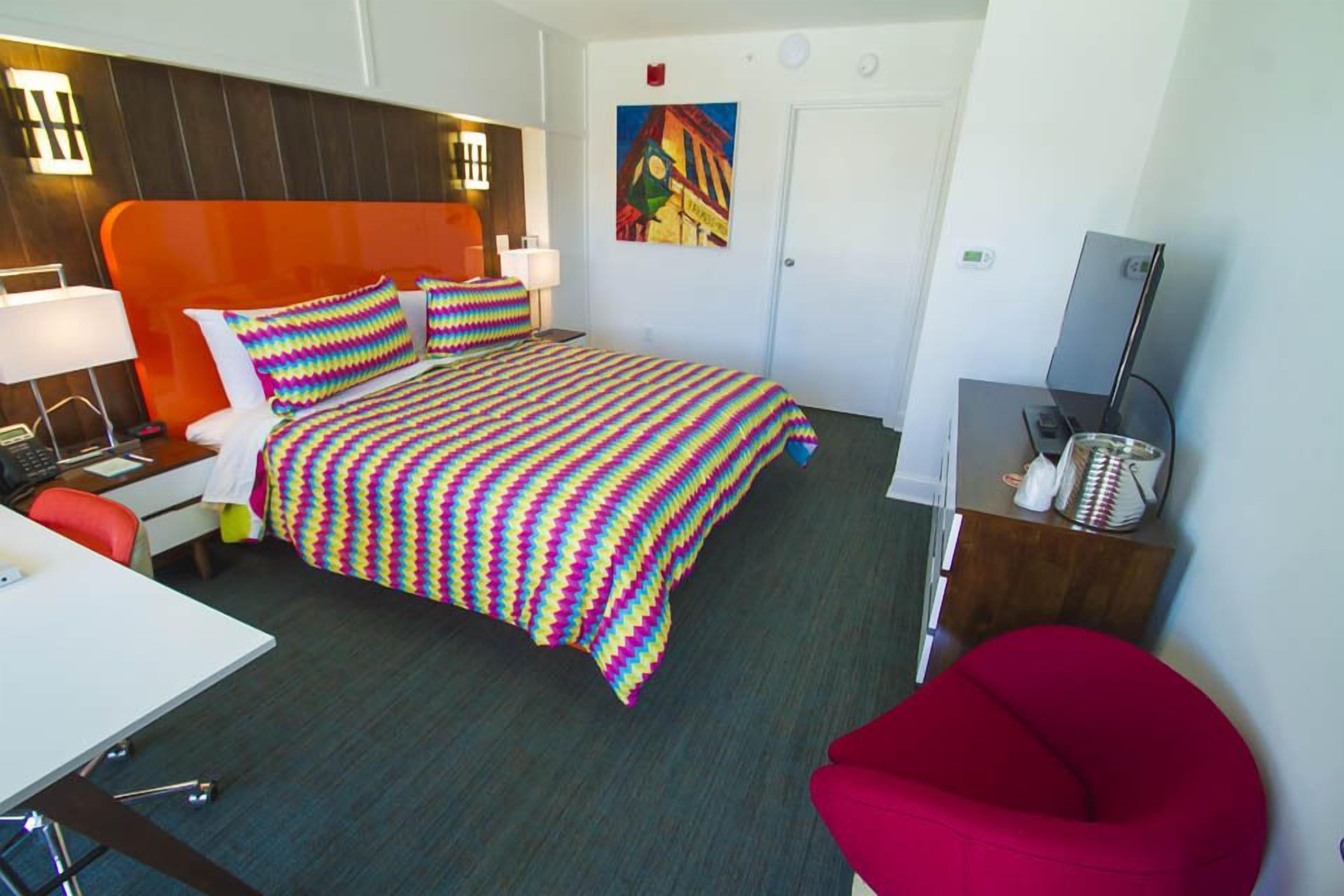 A hotel room with a bed, a desk, a chair, a TV, and a painting on the wall.