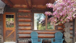 A log cabin with a wooden door marked "3", two blue Adirondack chairs, a small table, a window with a view of trees, and a hanging pink flowering plant.