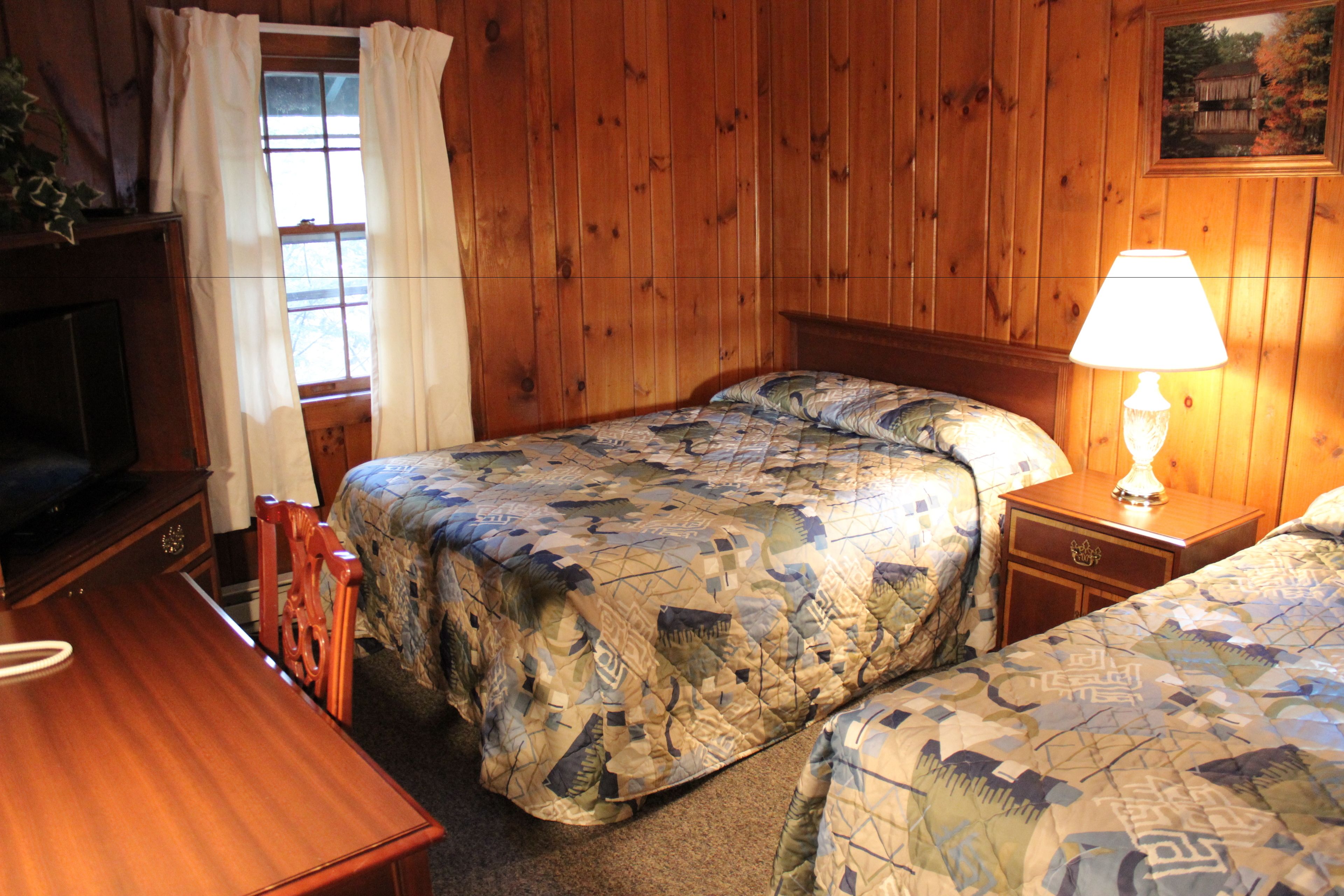 A room with wooden paneling, two beds with patterned quilts, a wooden nightstand with a lamp, a wooden desk, and a television.