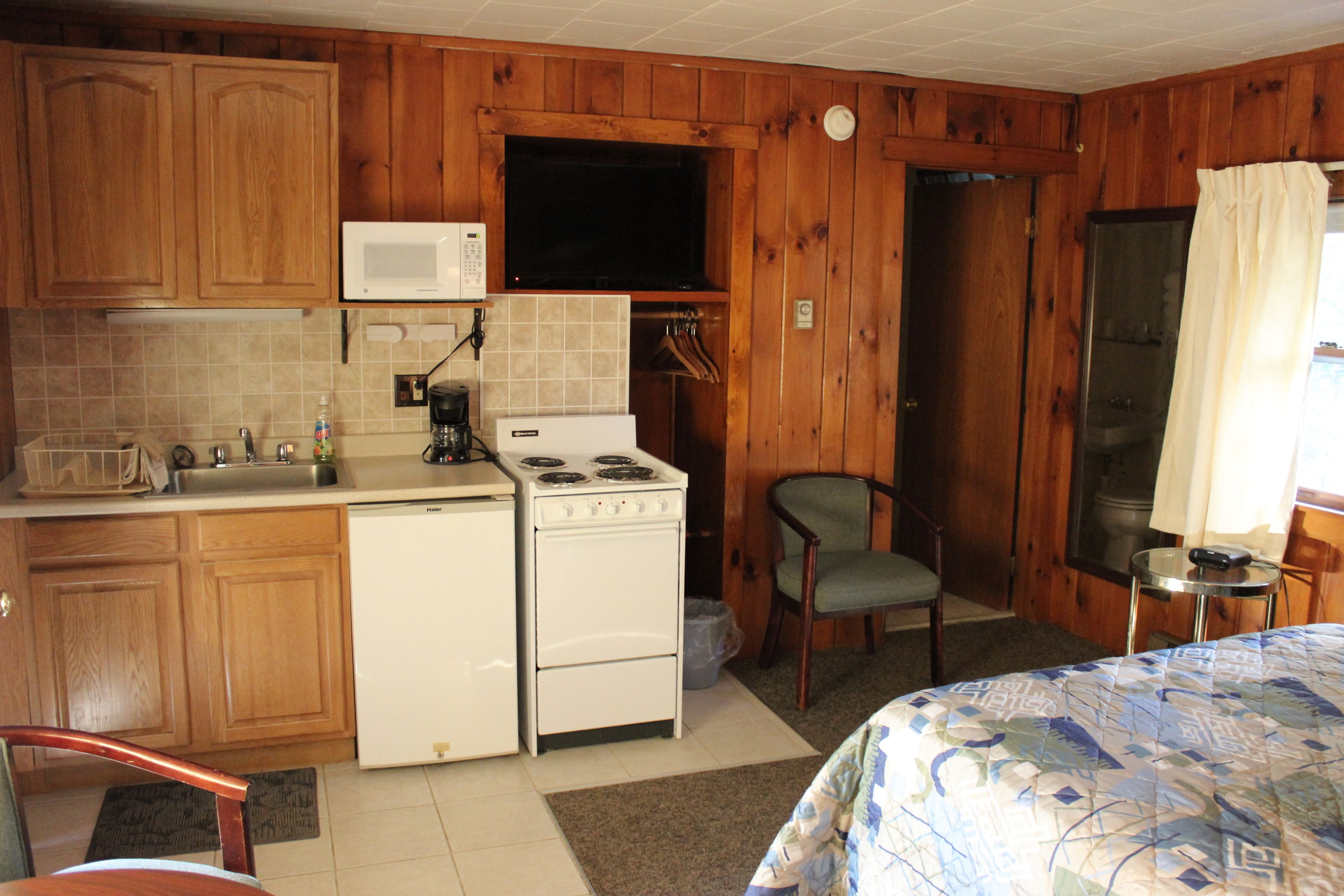 A compact kitchen with wooden cabinets, a microwave, a stove, and a sink. There is a chair and a small table with a remote control.